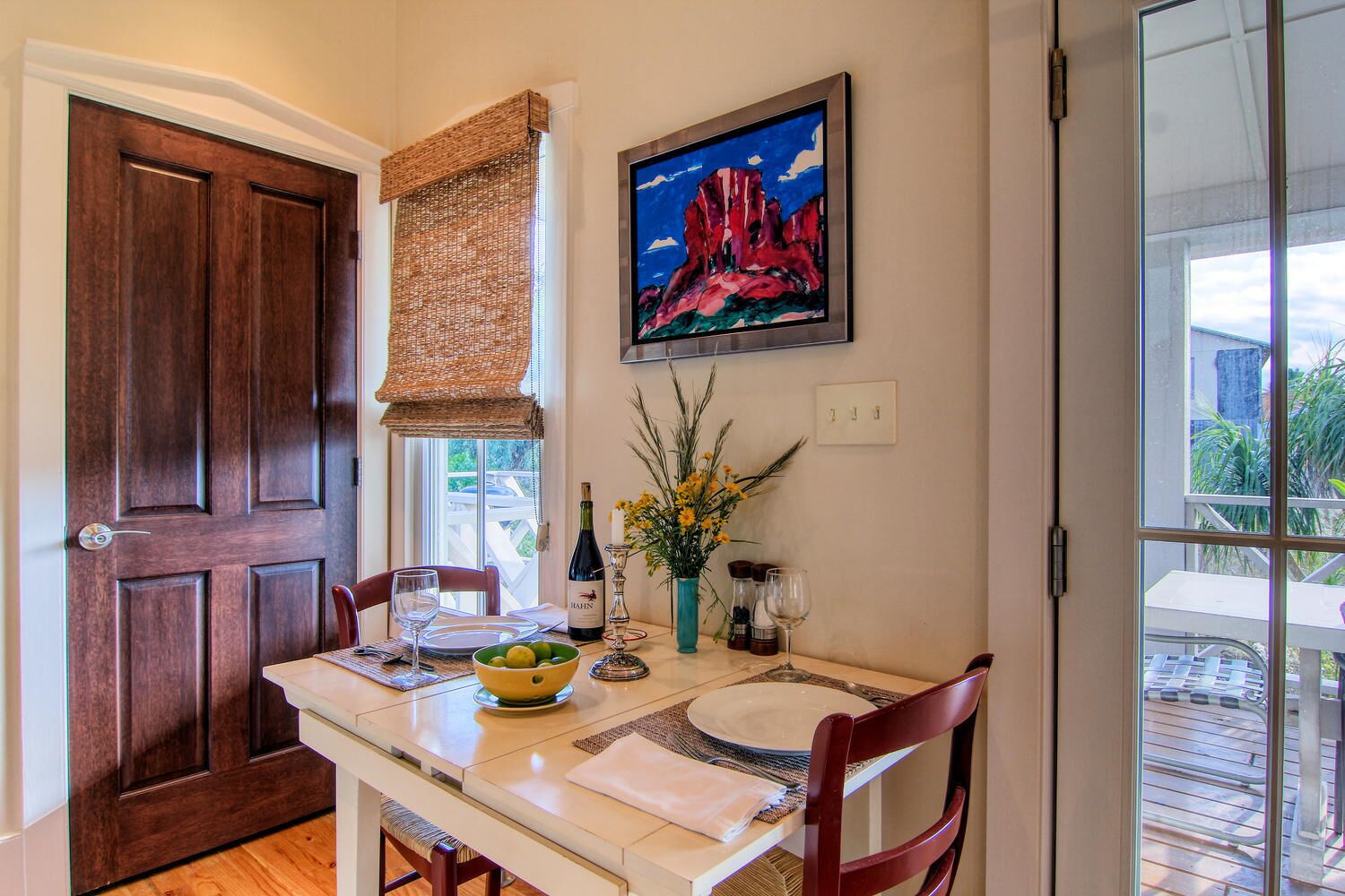 A dining area with a wooden door, a window with a woven blind, a painting of a mountain, and a table set with plates, glasses, and a bottle of wine.