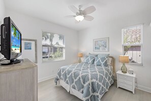 A bedroom with a bed, a TV on a stand, a ceiling fan, and two windows with blinds.