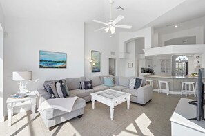 A spacious living room with a grey sofa, a coffee table, and a TV. There is a kitchen area with a bar and stools. - Relaxing Waterfront Home | Pool & Steps to Beach (Marco Island)