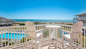 Un balcon avec des chaises blanches et une table donnant sur une piscine et une plage.