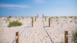 Un sentier de sable qui longe la plage et mène à l’océan, bordé de poteaux et de cordes en bois.