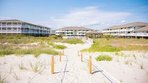 Un hôtel situé en bord de mer, doté de nombreux balcons, d’un chemin de sable menant à l’entrée et offrant une vue sur un ciel dégagé.