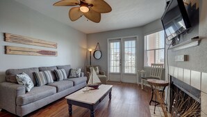 A living room with a grey sofa, a coffee table, a fireplace, and a ceiling fan. - Large Updated BEAUTIFUL 2BR, 2 BATH Condo! Walk to Beach, Pool & More! (Corpus Christi)