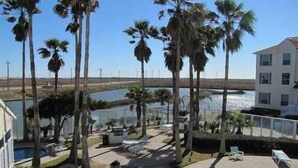 A poolside view with palm trees, a building, and a body of water. - Large Updated BEAUTIFUL 2BR, 2 BATH Condo! Walk to Beach, Pool & More! (Corpus Christi)