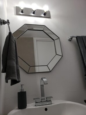 Un baño con un espejo hexagonal, un lavabo con grifo moderno y toallas colgadas en un perchero.