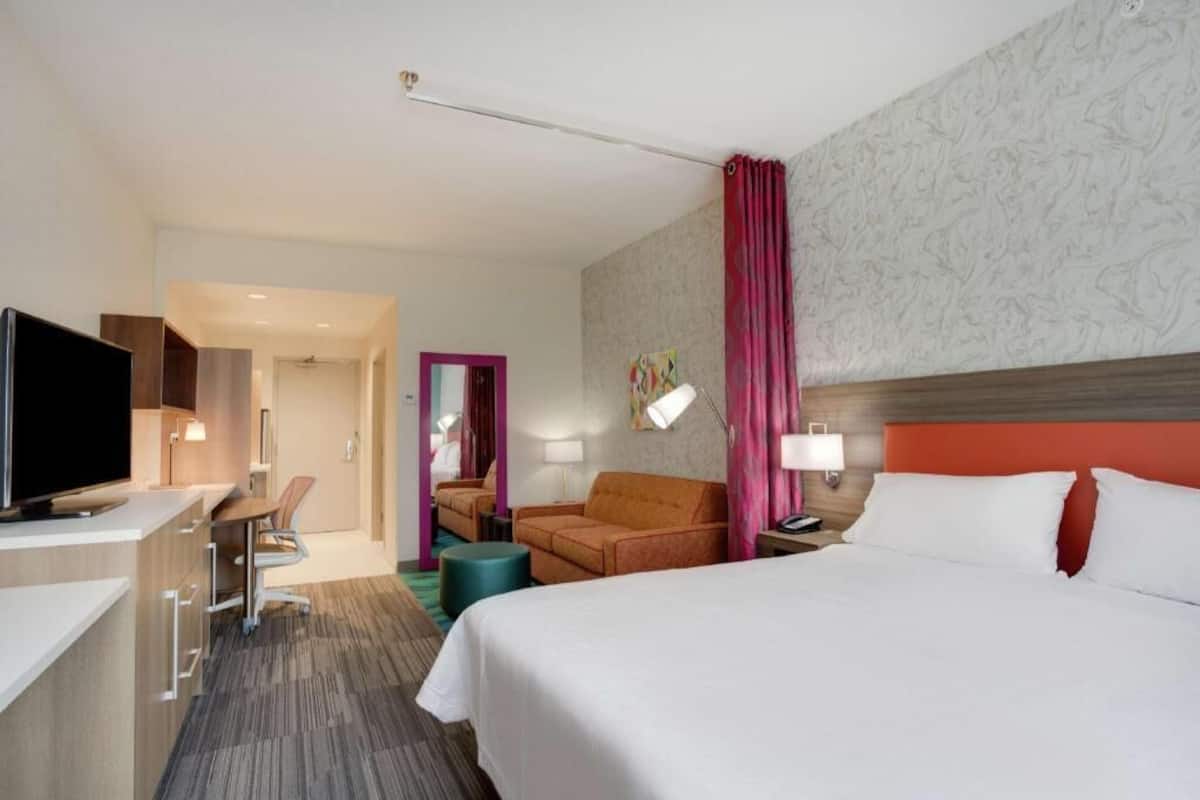 Une chambre d’hôtel comprenant un lit, un bureau, une télévision, une chaise, un canapé et une salle de bain, le tout visible à travers une porte ouverte.