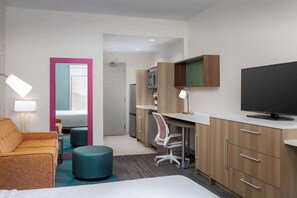 A hotel room with a bed, a desk with a TV, a chair, a sofa, and a bathroom visible through an open door. - Home2 Suites by Hilton Clermont (Clermont)