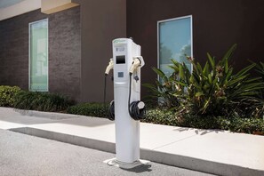 A white electric vehicle charging station with two cables, located on a sidewalk next to a building with large windows and a planter with green plants. - Home2 Suites by Hilton Clermont (Clermont)