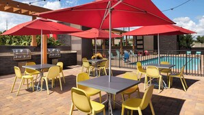 Une terrasse extérieure avec des chaises jaunes et des parasols rouges, un barbecue et une piscine.