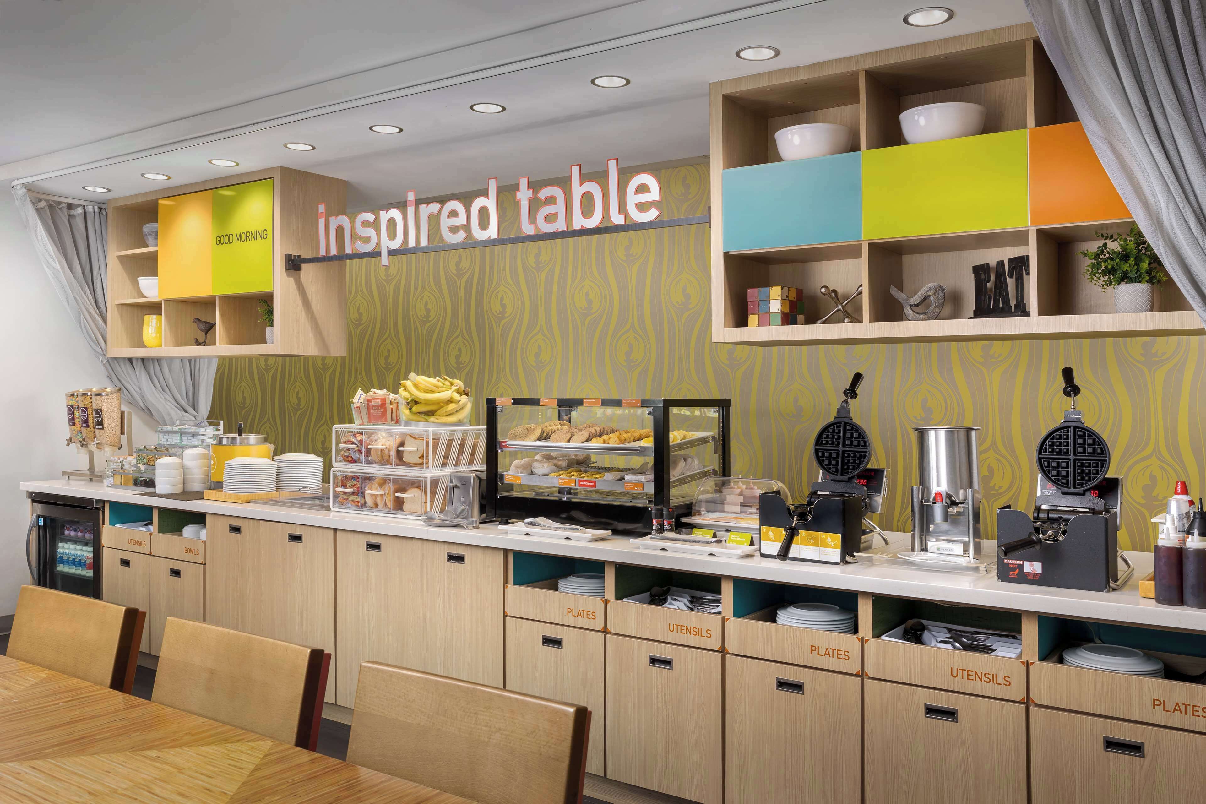 Una barra de desayuno con un letrero que dice "inspired table", que ofrece una variedad de alimentos para el desayuno, una cafetera y espacio para platos y cubiertos.