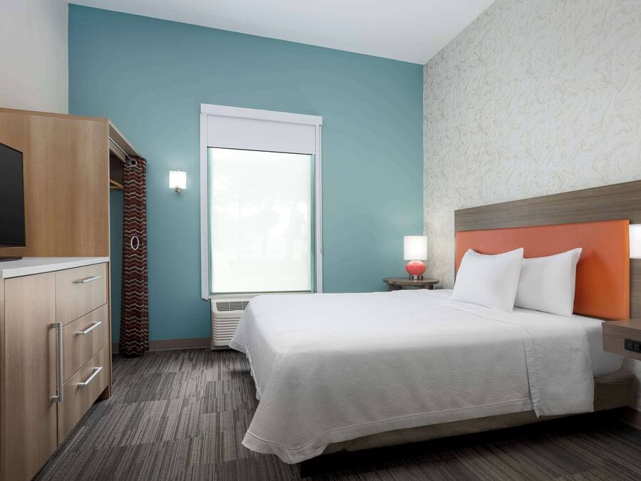 Una habitación de hotel con cama, televisor, armario, ventana y mesita de noche con lámpara.