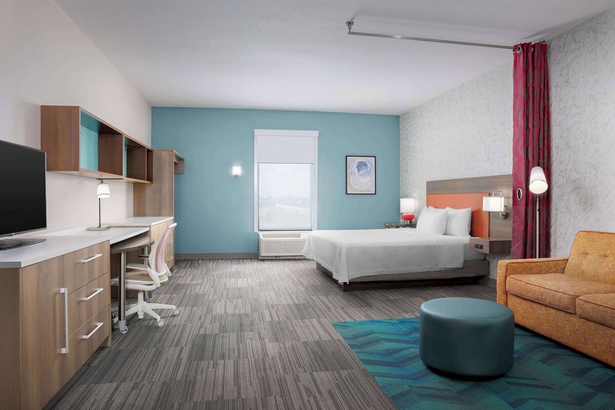 Une chambre d’hôtel avec un lit, un bureau avec un ordinateur, une chaise, un canapé et une fenêtre avec des rideaux.