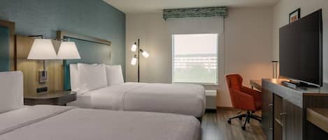 Habitación de hotel con dos camas, un escritorio con televisor, una silla y ventana con vista a edificios.