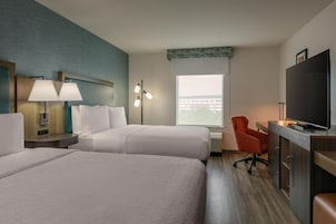 Quarto de hotel com duas camas, uma mesa de trabalho com televisão, uma cadeira e janela com vista para edifícios.