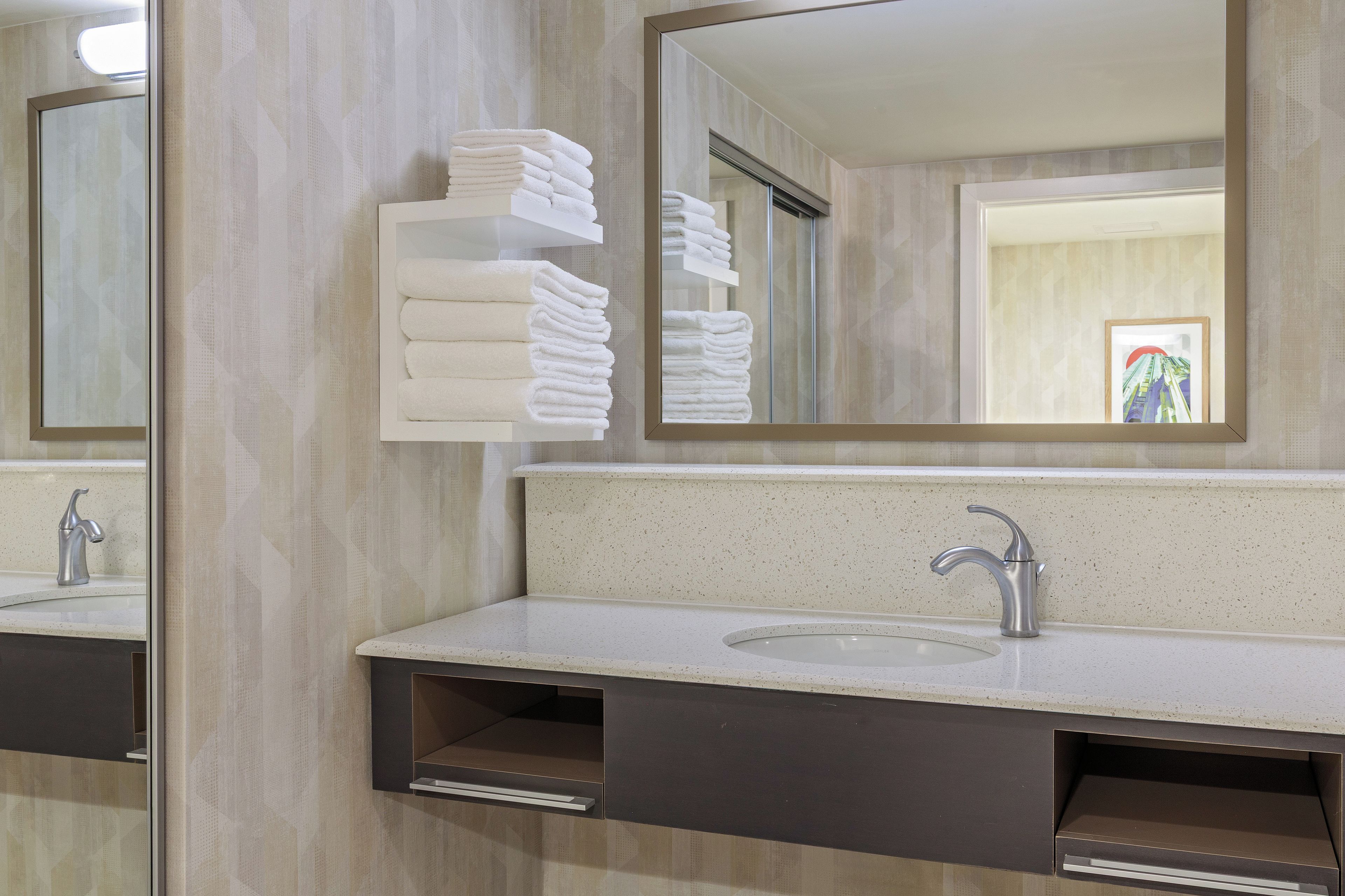 Un baño con lavamanos, espejo, toallero y puerta que ofrece vista a un edificio.
