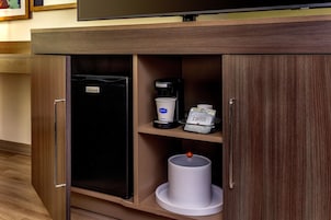 Un minibár de hotel con una cafetera, dispensador de cápsulas de café y un cesto de basura.