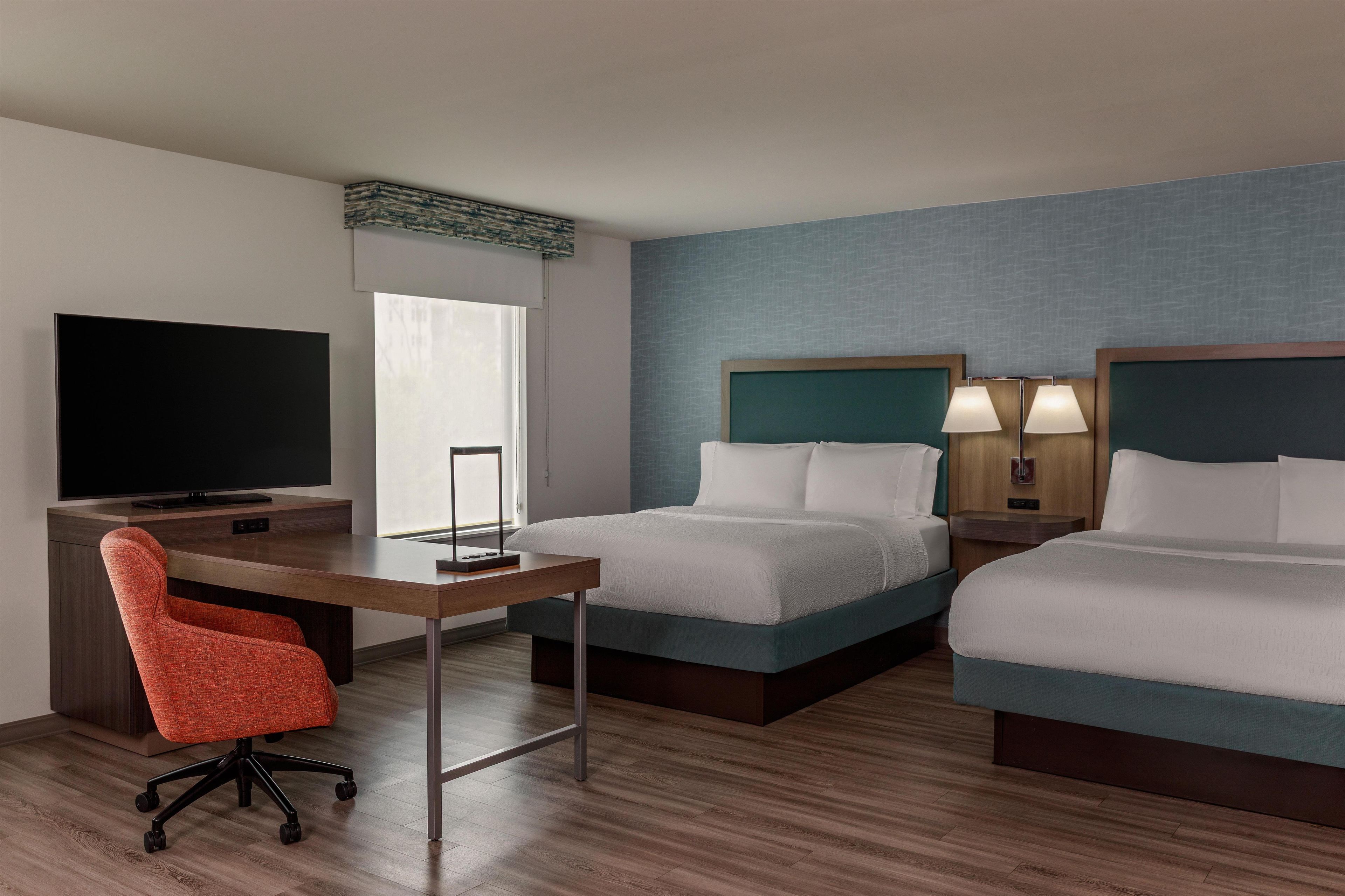 A hotel room with two beds, a desk with a TV, a chair, and a window.