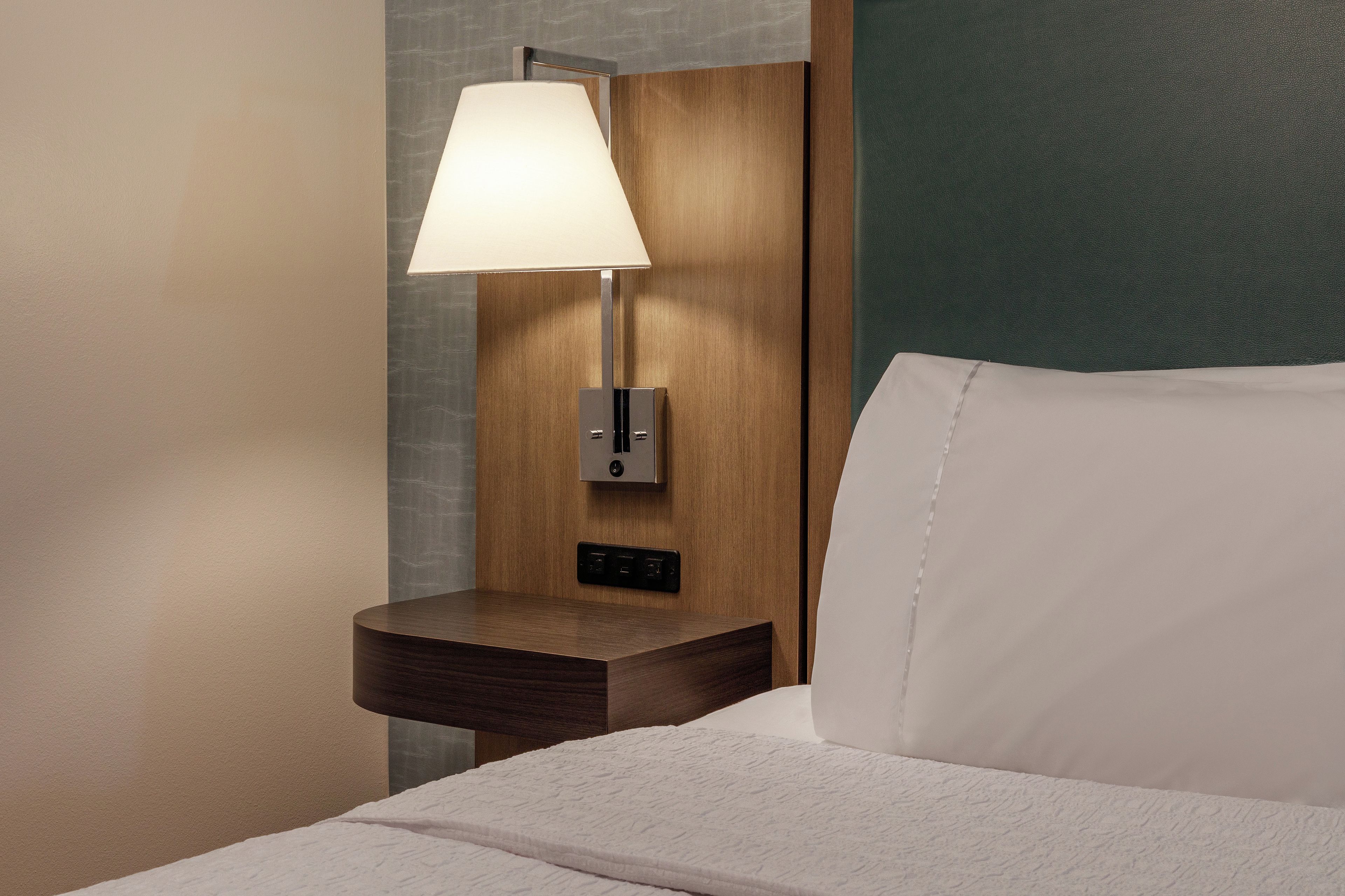A hotel room with a bed, a nightstand with a lamp, and a power outlet.