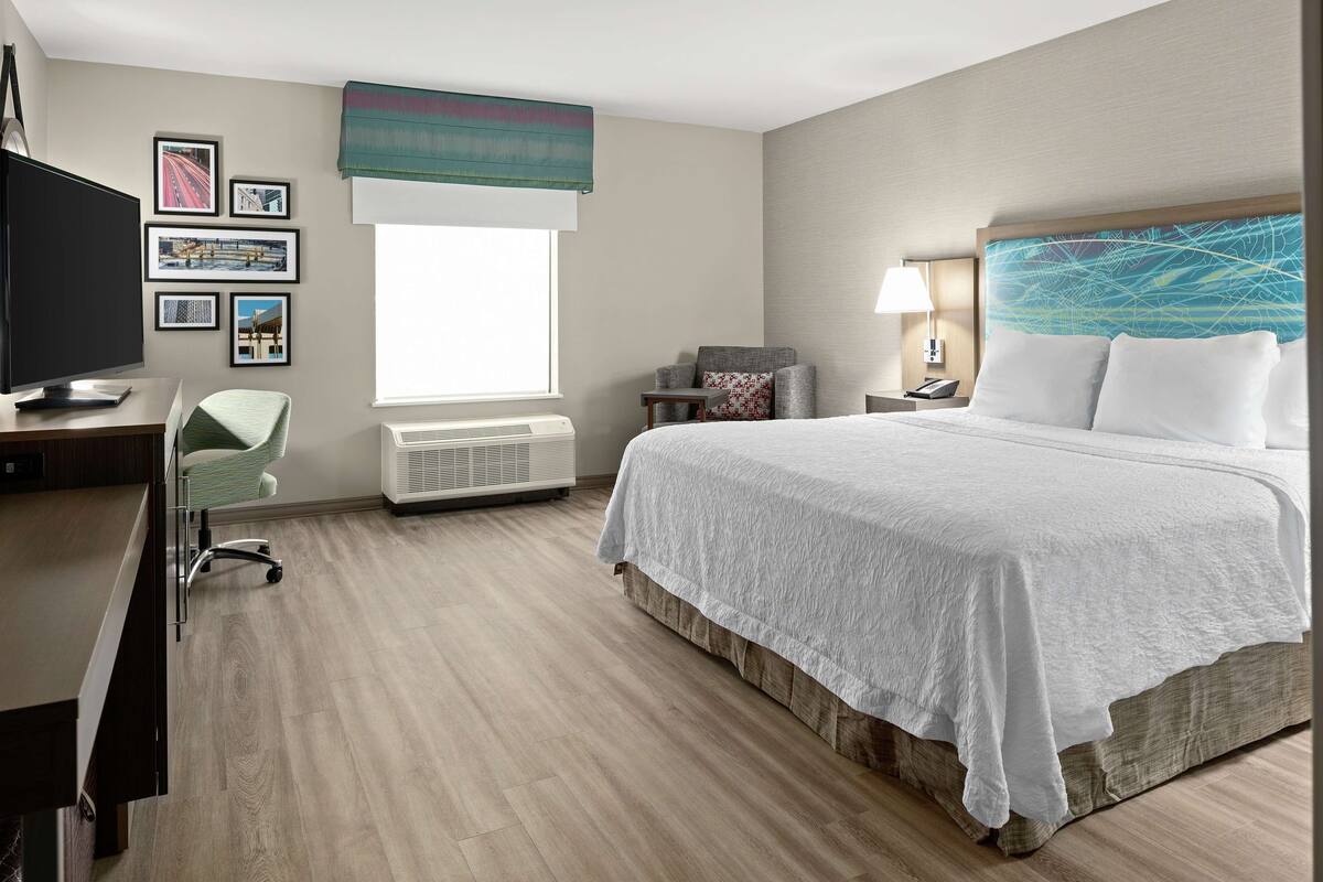 A hotel room with a large bed, a desk with a computer, a chair, a window with a view, and a wall with framed pictures.