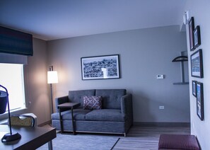 A living room with a grey sofa, a small side table, a lamp, and a framed cityscape picture on the wall. - Hampton Inn & Suites Cranberry Pittsburgh (Cranberry Township)