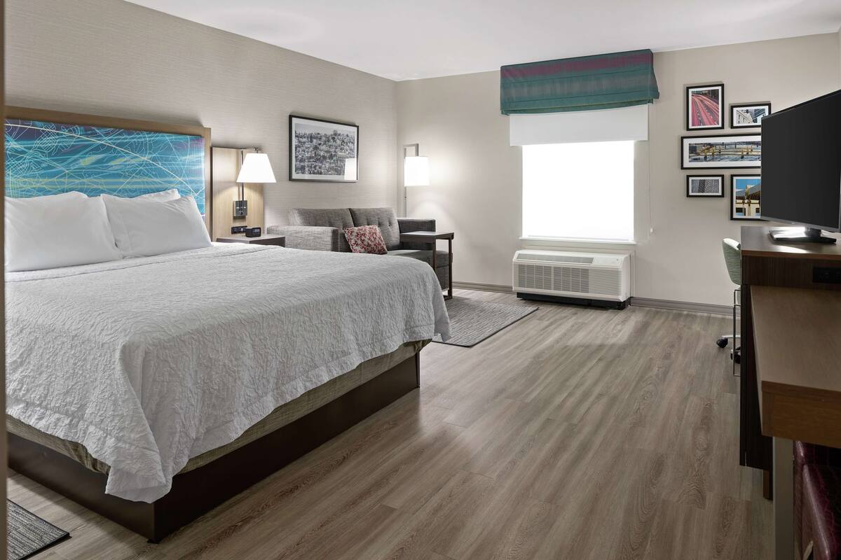A hotel room with a large bed, a desk with a flat-screen TV, a chair, a bedside table with a lamp, a wall-mounted air conditioner, and framed pictures on the wall.