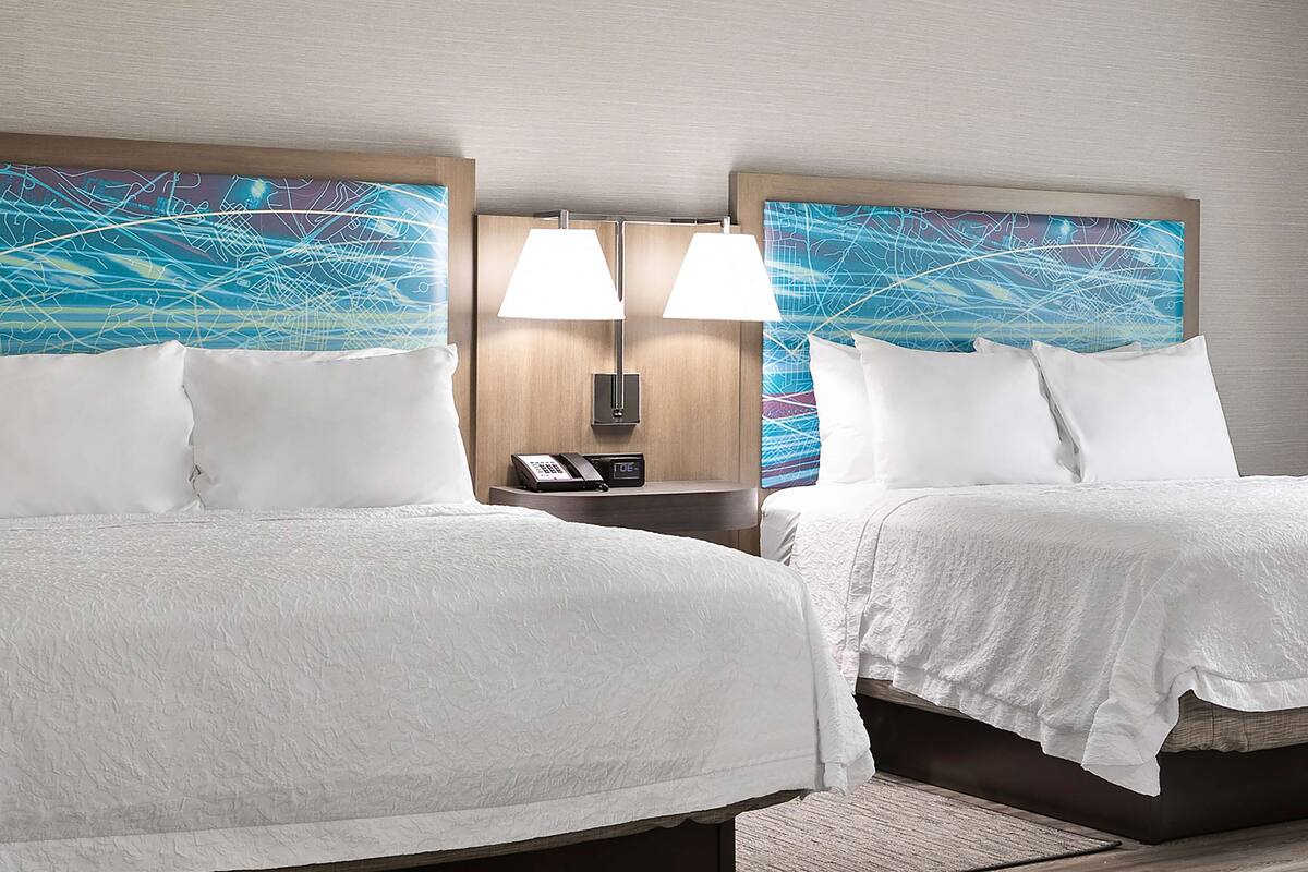 A hotel room with two beds, each with white bedding, a nightstand with a lamp, a phone, and a digital clock.