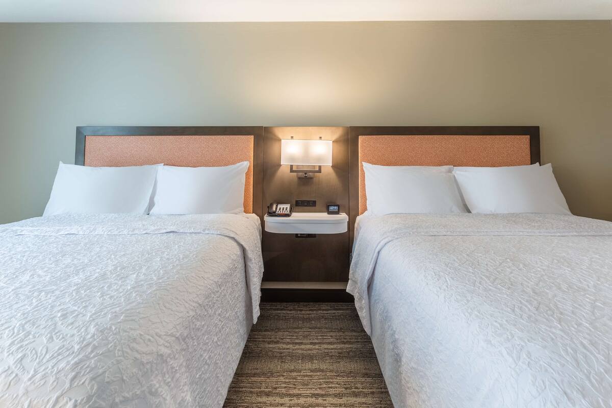 Quarto de hotel com duas camas, uma mesa de cabeceira, uma luminária na parede e um telefone.