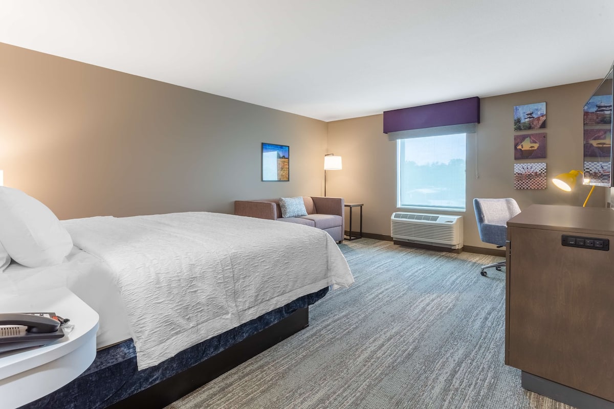 Quarto de hotel com uma cama grande, escrivaninha, cadeira, abajur e geladeira.