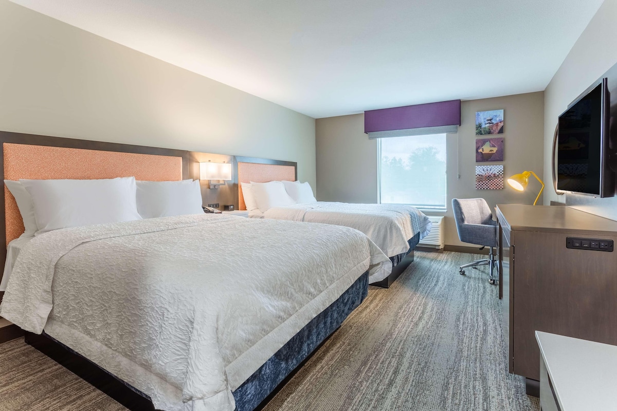 A hotel room with two beds, a desk, a chair, a TV, and artwork on the wall.