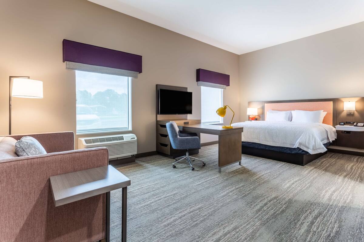 Quarto de hotel com cama, escrivaninha, cadeira, televisão e amplas janelas.