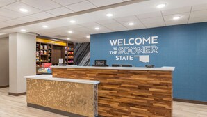 A hotel reception area with a wooden reception desk, a blue wall with "WELCOME TO THE SOONER STATE" signage, and a self-service kiosk. - Hampton Inn & Suites Guymon (Guymon)