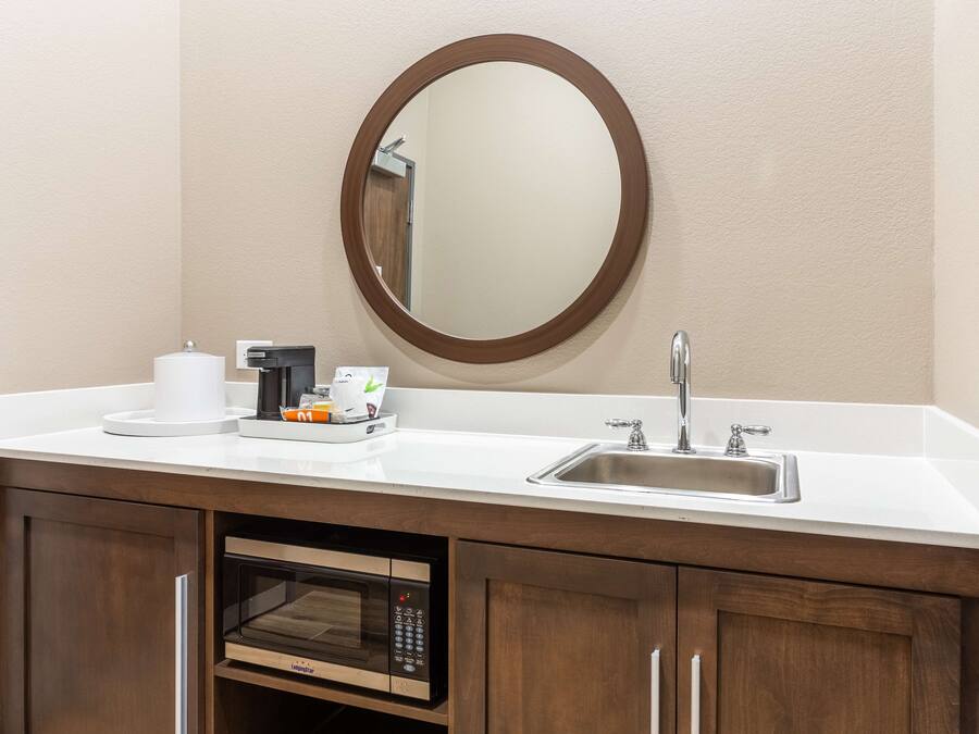 A modern bathroom with a white countertop, a sink, a mirror, a coffee maker, a microwave, and wooden cabinets.