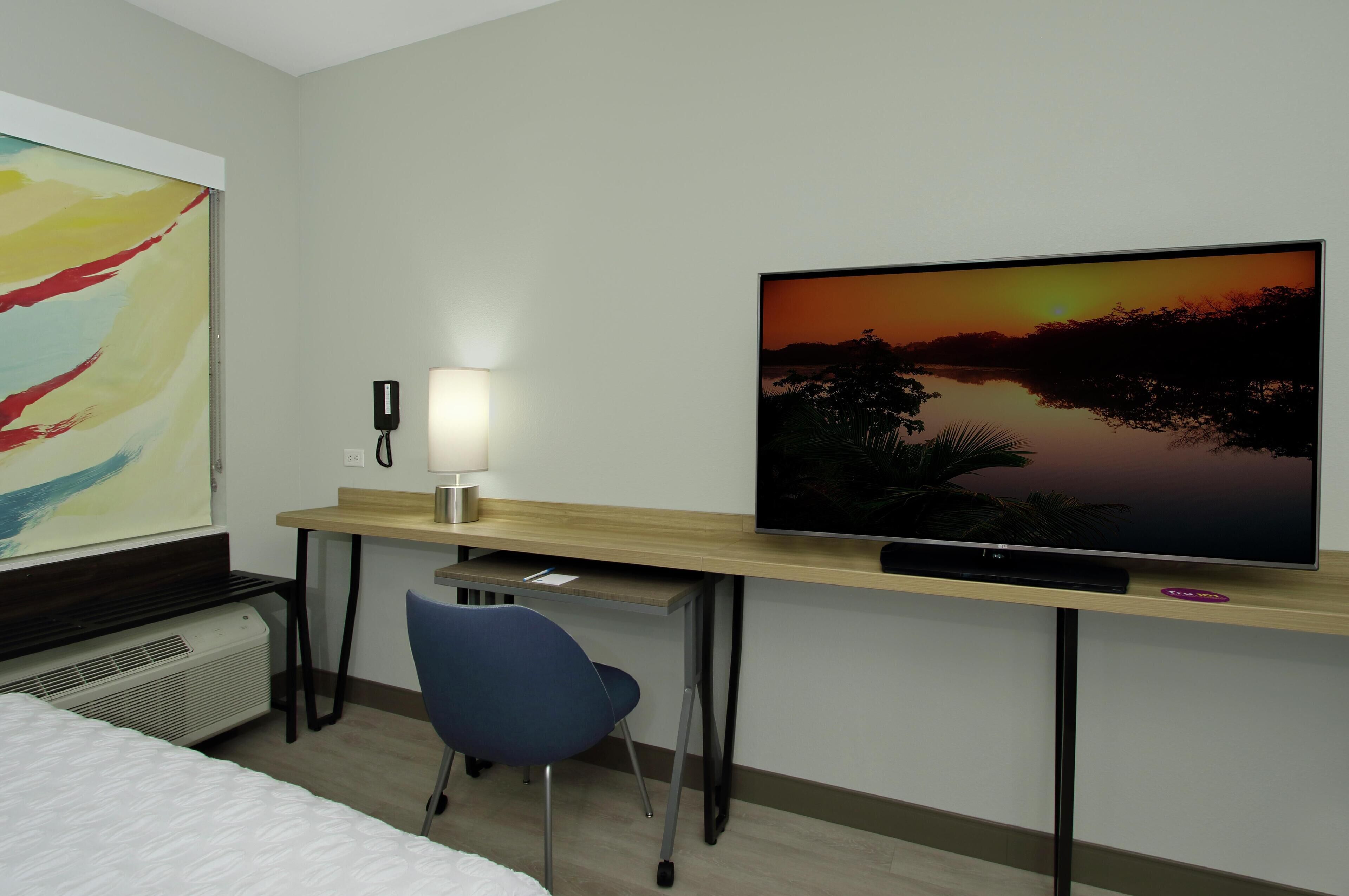 A hotel room with a bed, a desk with a computer, a TV displaying a sunset, a lamp, and a phone.