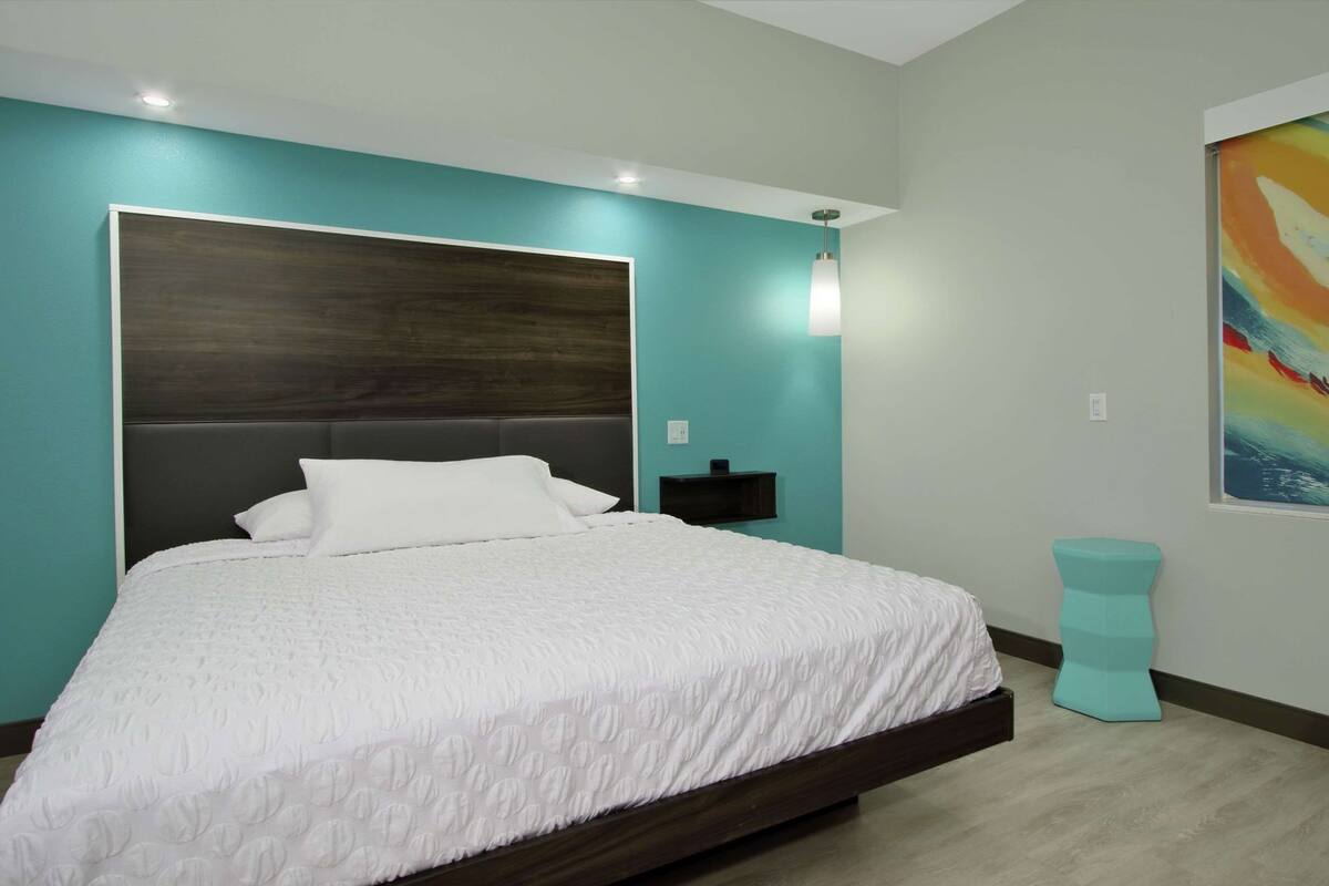A modern bedroom with a large bed, a nightstand, a teal accent wall, and a colorful abstract painting.
