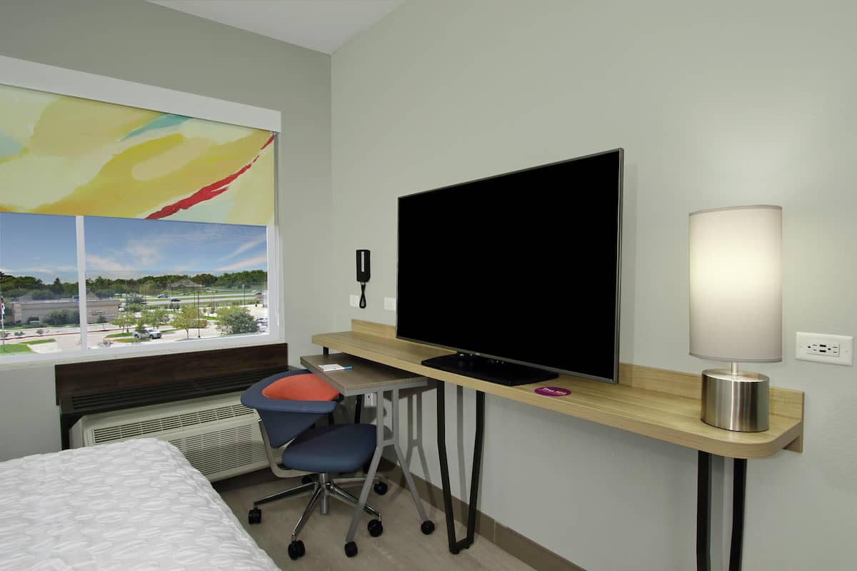 Ett hotellrum med en säng, ett skrivbord med en tv, en stol och ett fönster med utsikt över staden.