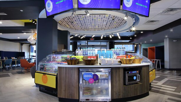 Zona de recepción de hotel con diseño moderno, que incluye un mostrador curvo, varias pantallas mostrando el nombre del hotel y una variedad de servicios como un refrigerador y una máquina expendedora.