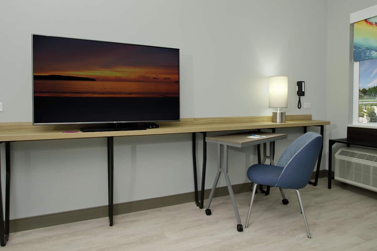 Ett hotellrum med en stor platt-TV på ett skrivbord i trä, en skrivbordslampa, en stol och ett fönster med utsikt över ett landskap.