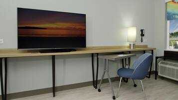 A hotel room with a large flat-screen TV on a wooden desk, a desk lamp, a chair, and a window with a view of a landscape.