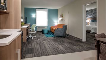A hotel room with a kitchenette, a seating area with a sofa and armchair, a bed with a headboard, and a window with blinds.