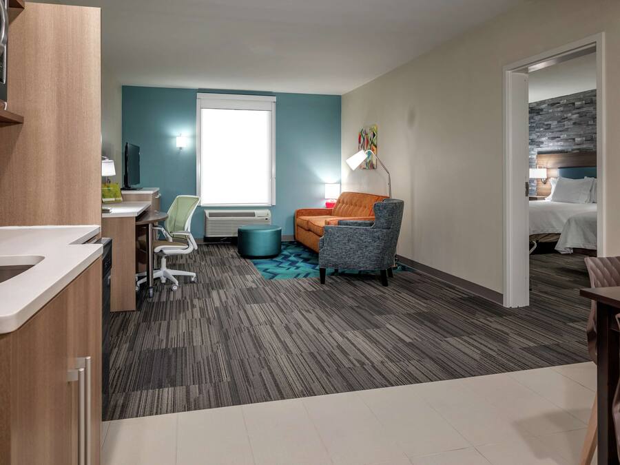 Habitación de hotel con kitchenette, zona de estar con sofá y sillón, cama con cabecera y ventana con persianas.