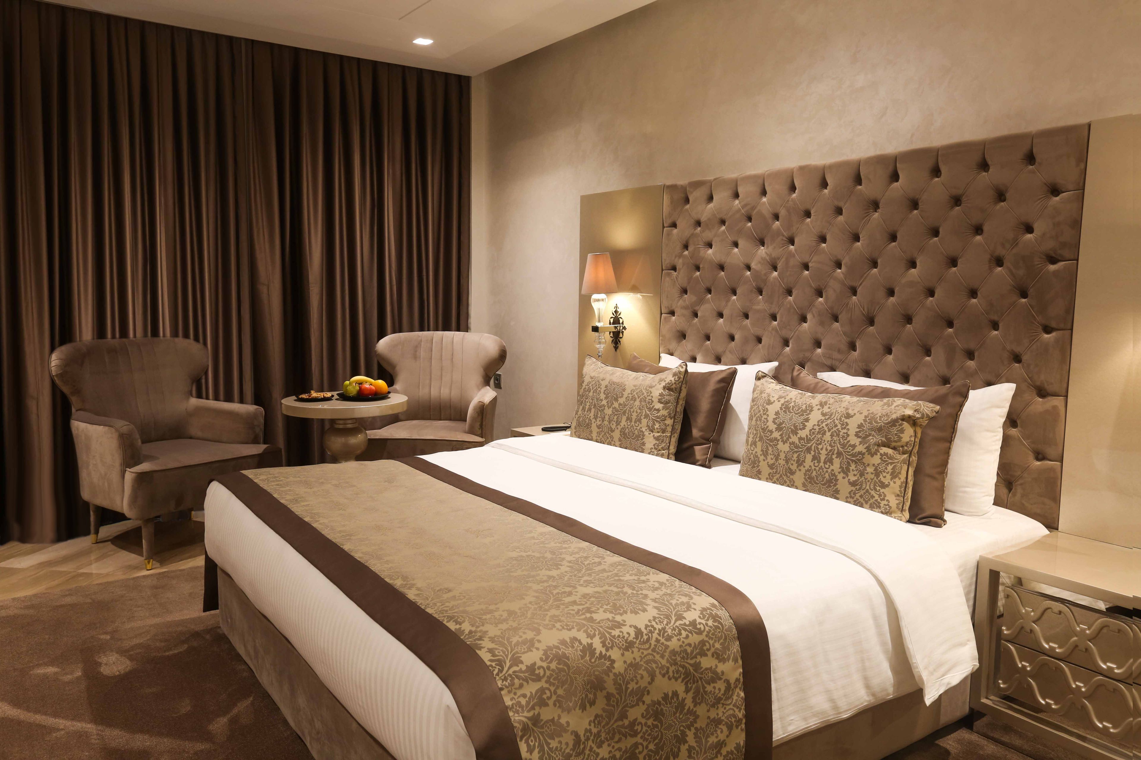 A hotel room with a large bed, two armchairs, a small round table with fruit, and a wall-mounted lamp.