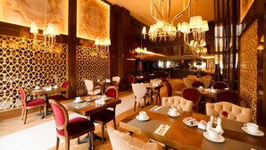 Een goed verlicht restaurant met meerdere eettafels en stoelen, een kroonluchter en een decoratieve wandbekleding.