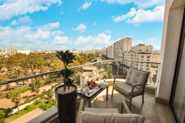 A balcony with a view of a cityscape, a potted plant, and a table with breakfast items.