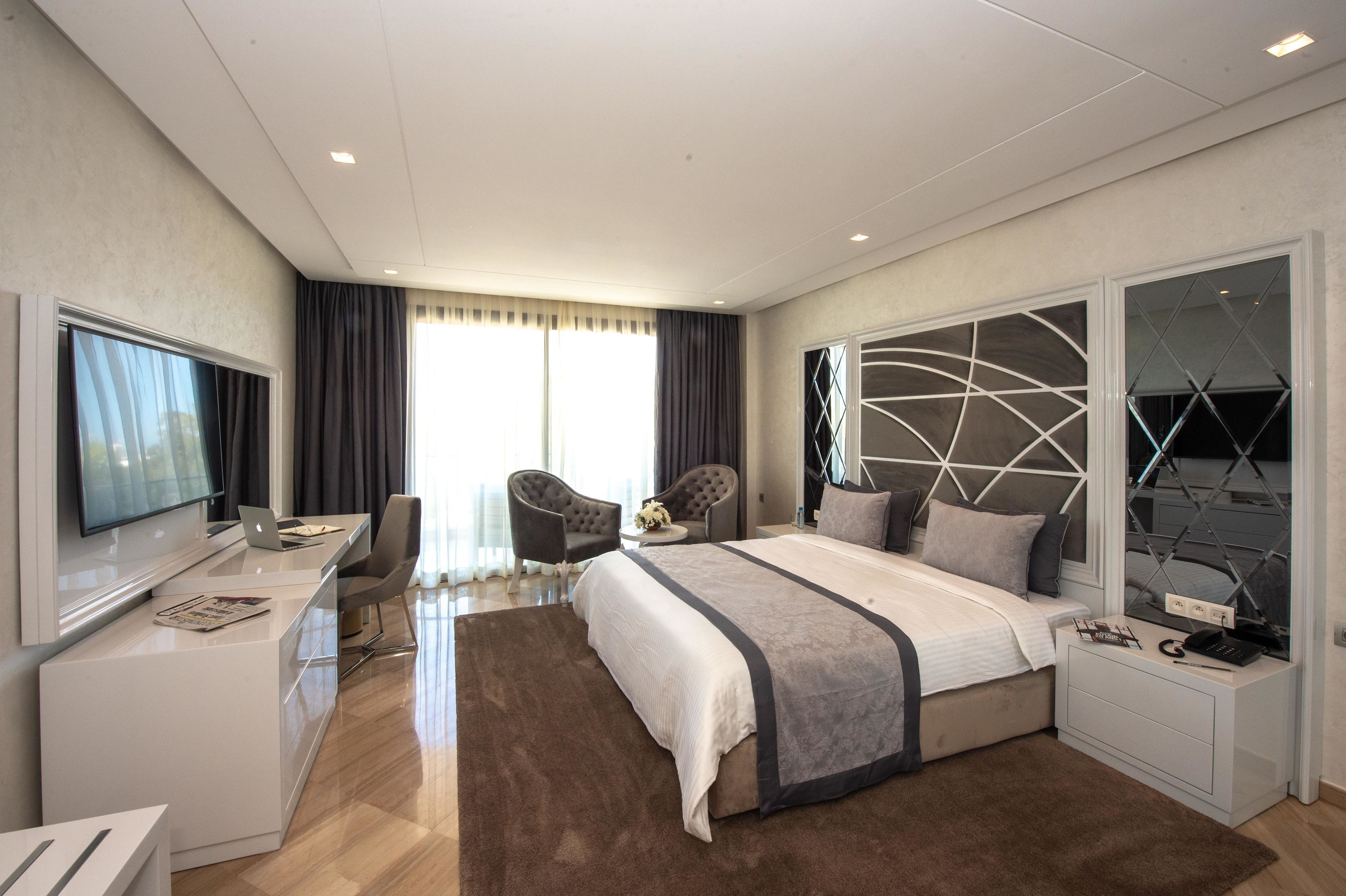 A modern hotel room with a large bed, a desk with a laptop, a TV, and a seating area with a chair and a small table.