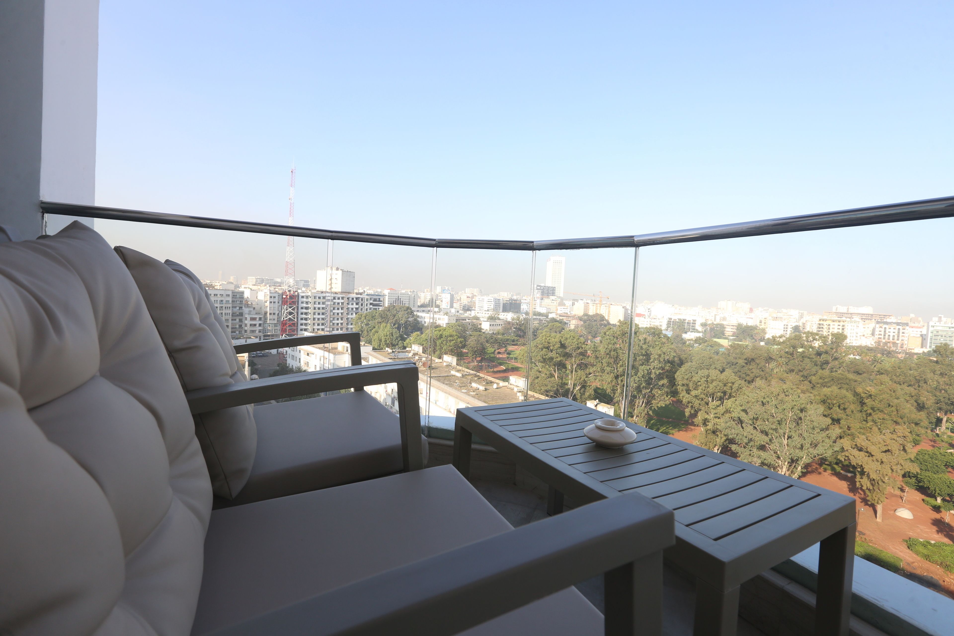 A balcony with a cushioned seating area and a small table, overlooking a cityscape with buildings and trees.