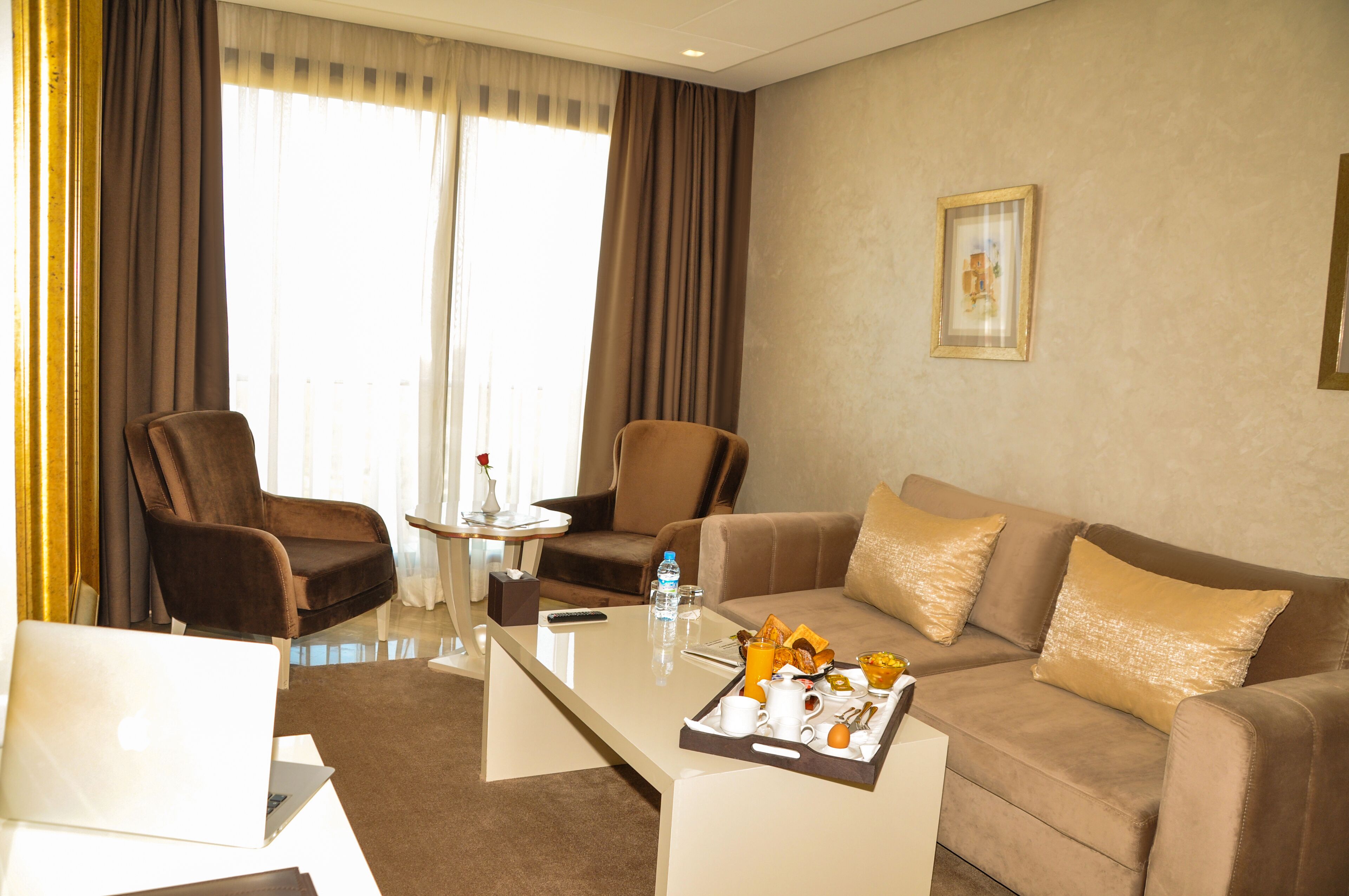 Ein Hotelzimmer mit einem braunen Sofa, zwei Sesseln, einem Glastisch und einem Laptop auf einem weißen Schreibtisch.