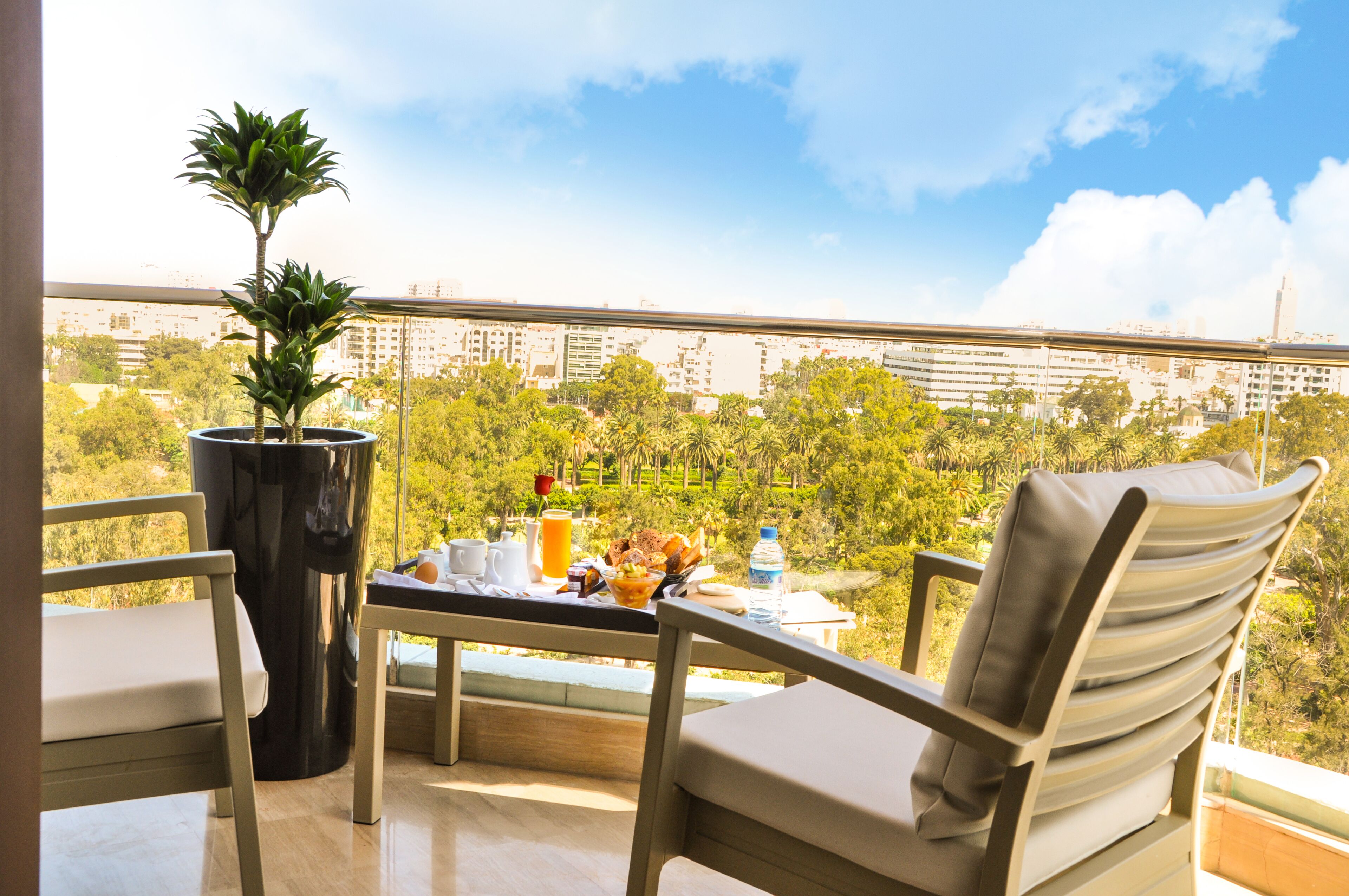 A balcony with a table set for breakfast, a potted plant, and a view of a cityscape.