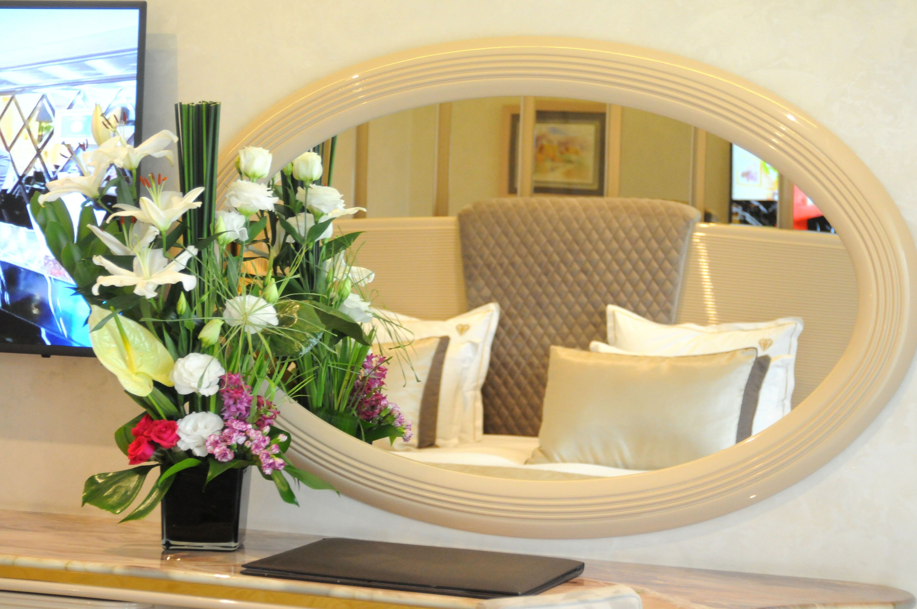 Una habitación con un espejo ovalado grande que refleja una cama con ropa de cama blanca y marrón, un florero con flores sobre una mesa, y un televisor en la pared.