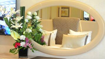 Una habitación con un espejo ovalado grande que refleja una cama con ropa de cama blanca y marrón, un florero con flores sobre una mesa, y un televisor en la pared.