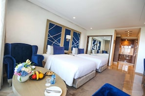 A hotel room with two beds, a dining table with fruit and a vase of flowers, and a large mirror on the wall.